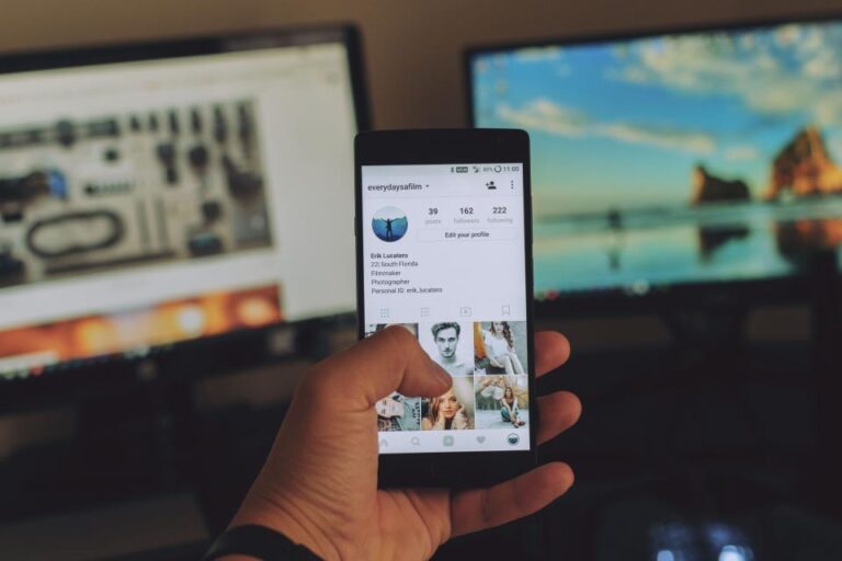 Hand holding a phone showing a chat conversation at a desk with monitors